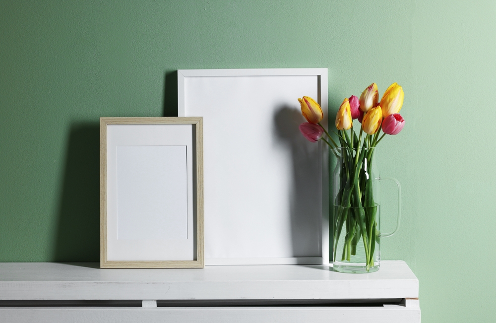 Two empty picture frames on a white shelf next to some tulip flowers, with a green wall in the background.
